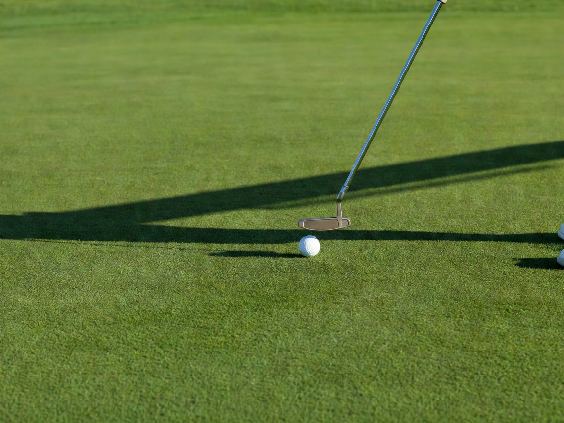 Close view of a putter meeting the ball on a smooth green, the shaft leaning slightly forward and the player's shadow across the turf